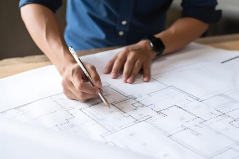 Osoba trzymająca ołówek nad dużym arkuszem papieru z planami budynku leżącym na stole