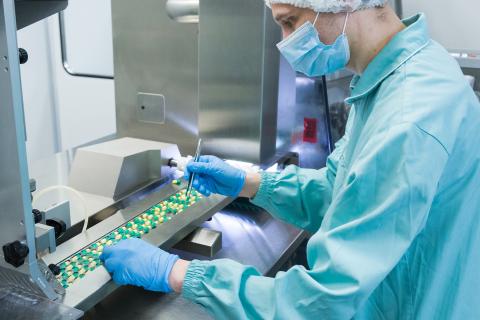 Mężczyzna w odzieży ochronnej pracuje przy taśmie produkcyjnej, na której znajdują się kapsułki z lekami