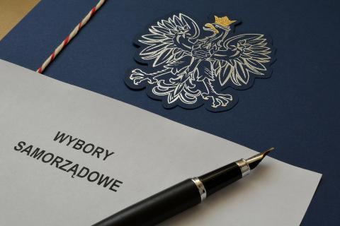 Pióro i kartka z napisem "Wybory samorządowe" ułożone na teczce z godłem Polski - orłem w koronie