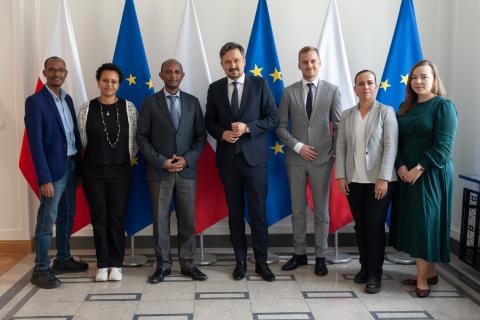 Siedem osób pozuje na stojąco do wspólnego zdjęcia na tle flag Polski i UE