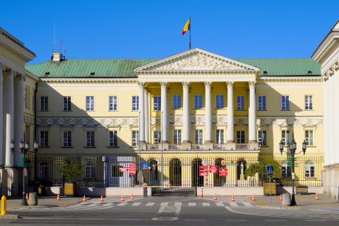 Zabytkowy budynek w styli klasycystycznym z portykiem kolumnowym, przed budynkiem na ogrodzeniu czerwone tablice z nazwami urzędów