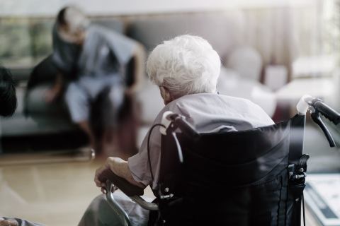Starsza osoba siedzi na wózku zwróconym tyłem do kadru. W tle rozmyta sylwetka drugiej osoby wstającej z kanapy 