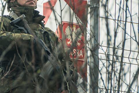 Żołnierz trzymający karabin stoi przy biało-czerwonym słupie granicznym z herbem Polski i napisem "Polska"