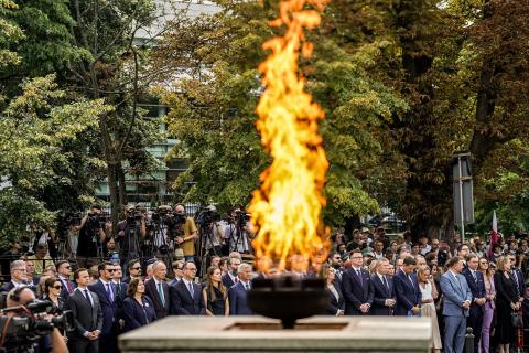 Kilkadziesiąt osób stoi oddając hołd Powstańcom Warszawskim, na pierwszym planie płonący znicz