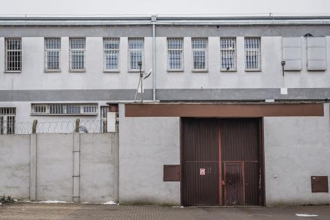 Stalowa brama zakładu karnego w wysokim murze zakończonym drutem kolczastym. Za bramą widoczny jest budynek z okratowanymi oknami