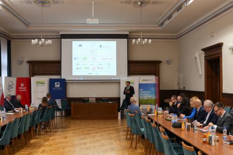 Osoba przemawia na stojąco na sali konferencyjnej, a osoby siedzące za stołami ustawionymi w podkowę słuchają
