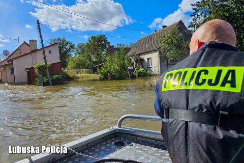 osoba z napisem policja siedzi tyłem na łodzi, w tle zalane wodą domy