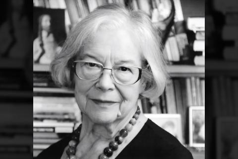 Czarno-biały portret Krystyny Mrugalskiej - kobieta w okularach i z koralami na szyi na tle regału z książkami i zdjęciami