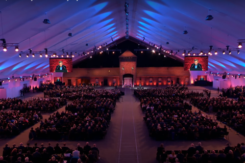 Duży namiot rozstawiony nad bramą obozu Auschwitz, w którym siedzi bardzo wiele osób uczestniczących w uroczystościach. 
