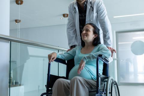 Uśmiechająca się kobieta w ciąży siedzi na wózku, który jest prowadzony przez mężczyznę w białym fartuchu