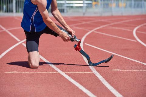 Mężczyzna w sportowym ubraniu przygotowuje się do biegu, klęcząc na bieżni i poprawiając protezę nogi