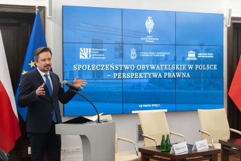 Osoba przemawia za mównicą na tle flag Polski i UE i wyświetlacza z napisem "Społeczeństwo obywatelskie w Polsce - perspektywa prawna"