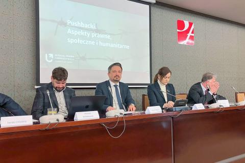 Cztery osoby, w tym Marcin Wiącek, siedzą przy prezydialnym stole. Za nimi wyświetla się tytułowy slajd prezentacji z napisem "Pushbacki. Aspekty prawne, społeczne i humanitarne"