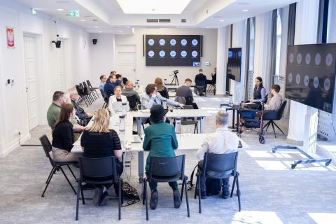 Kilkadziesiąt osób siedzi przy stołach na sali konferencyjnej, na dużych ekranach widoczni uczestnicy spotkania łączący się za pomocą oprogramowania do komunikowania się na odległość