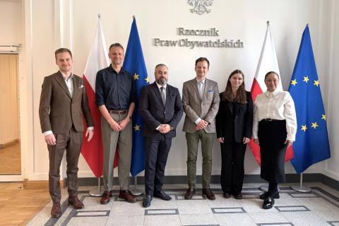 Sześć osób pozuje na stojąco do wspólnego zdjęcia na tle flag Polski i EU oraz napisu "Rzecznik Praw Obywatelskich" na ścianie