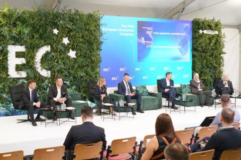 Siedem osób siedzi na fotelach na podwyższeniu na sali konferencyjnej podczas kongresu