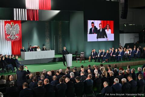 Kilkadziesiąt osób siedzących w dużej hali słucha wystąpienia osoby mówiącej za mównicą, na ścianie w tyle duże godło Polski - biały orzeł w koronie na czerwonym polu