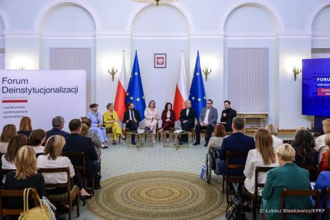 Osiem osób siedzi na krzesłach na tle flag Polski i UE, kilkadziesiąt osób na sali przysłuchuje się