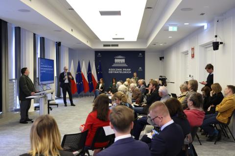 Widok ogólny sali, w której odbywa się debata. Przy mównicy w środkowej części sali stoi mężczyzna. Obok niego stoi drugi mężczyzna, który wypowiada się do mikrofonu, gestykulując. Wokół nich, na krzesłach ustawionych w kilku rzędach w półokrąg, siedzą inni uczestnicy wydarzenia.