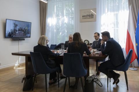 Pięć osób siedzi przy stole w sali konferencyjnej i uczestniczy w telekonferencji z trzema innymi osobami, wyświetlanymi na ekranie znajdującym się na ścianie.