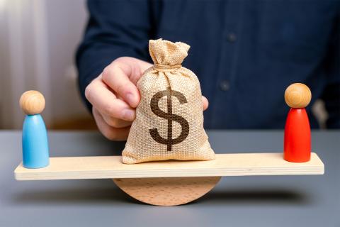 Ilustracja przedstawiająca dwie figurki stojące na przeciwległych końcach równoważni, oraz duży worek z symbolem dolara stojący na jej środku.