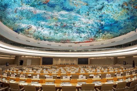 Sala obrad Rady Praw Człowieka ONZ. Kilka szeregów długich stołów jest ustawionych w okręgu wokół stołu prezydium. Nad stołami widać rzeźbę stanowiącą sufit pomieszczenia - kompozycję olbrzymiej ilości kolorowych, zastygniętych stalaktytów zwisających z sufitu.