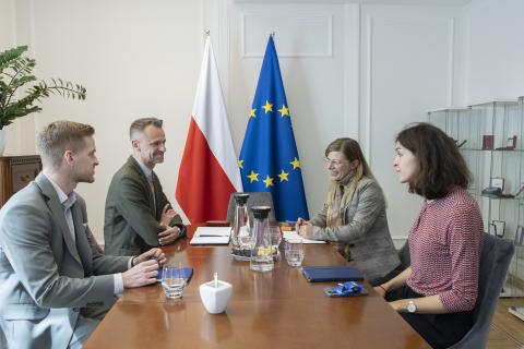 Cztery osoby siedzą po dwóch stronach stołu i rozmawiają, za stołem flagi Polski i UE