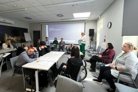 Kilkanaście osób siedzi przy stołach na sali, w tle na ekranie plansza z napisem "Prawo do dostępności"