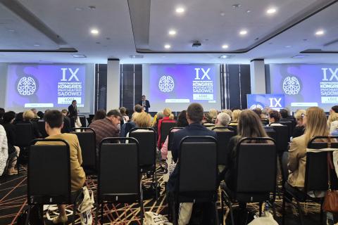 Kilkadziesiąt osób na dużej sali konferencyjnej siedzi na krzesłach i słucha wystąpienia osoby mówiącej z podwyższenia, w tle na trzech wyświetlaczach grafika z napisem "IX Międzynarodowy Kongres Psychiatrii Sądowej - Łódź 9-10 maja 2025
