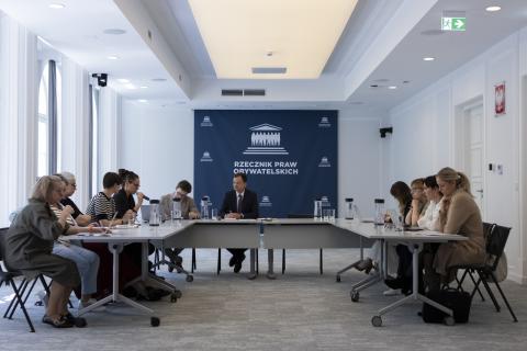 Kilkanaście osób siedzi przy stołach ustawionych w podkowę, za stołami przy ścianie rząd flag Polski i UE