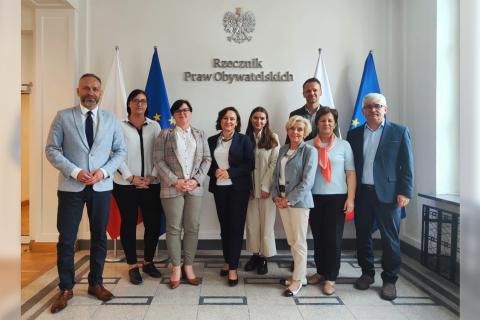 Dziewięć osób pozuje do zdjęcia stojąc przy flagach Polski i Unii Europejskiej, pod umieszczonym na ścianie napisem "Rzecznik Praw Obywatelskich" i symbolem orła