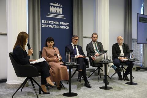 Pięć osób siedzi na ustawionych w rzędzie krzesłach. Jedna z nich wypowiada się w stronę innych, trzymając mikrofon.