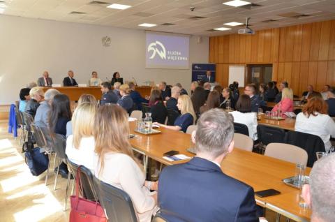 Kilkudziesięciu uczestników Kongresu siedzi przy długich stołach w sali konferencyjnej. W szczycie sali znajduje się prezydium, przy którym siedzą cztery osoby, oraz podium, przy którym stoi przemawiająca osoba. 
