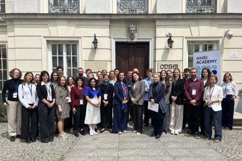 Grupa kilkudziesięciu osób stoi przed wejściem do budynku z napisem ODIHR