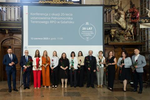 Kilkanaście osób pozuje na stojąco, trzymając czerwone róże, do wspólnego zdjęcia w zabytkowej, bogato zdobionej sali w Dworze Artusa