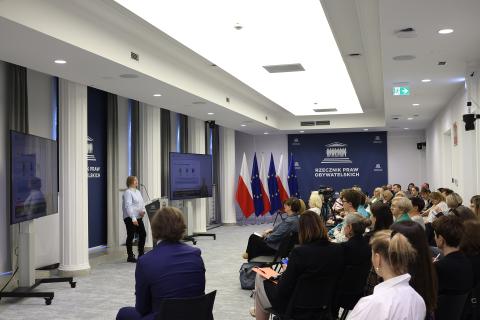 Widok ogólny sali konferencyjnej. Przy podium na środku sali stoi wypowiadająca się osoba. Przed nią na widowni siedzi kilkadziesiąt osób