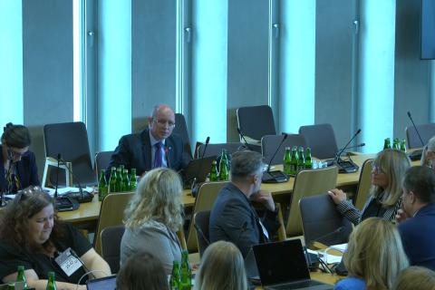 Kilkadziesiąt osób siedzi na sali konferencyjnej słucha osoby, która mówi do mikrofonu