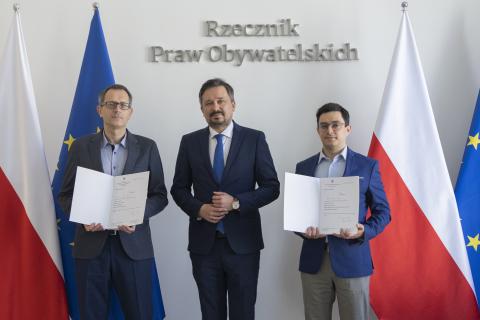 Tomasz Bzukała, Marcin Wiącek i Michał Szwast pozują do zdjęcia na tle flag Polski i Unii Europejskiej i napisu "Rzecznik Praw Obywatelskich". Tomasz Bzukała i Michał Szwast trzymają w dłoniach otwarte teczki z powołaniami na stanowiska.