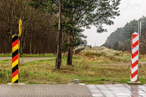 Widok na pas graniczny na granicy Polski i Niemiec - wykarczowany teren o szerokości kilkudziesięciu metrów w lesie. Na pierwszym planie widać chodnik, przy którym stoją dwa słupy graniczne - polski, biało-czerwony, oraz niemiecki, żółto-czarno-czerwony.