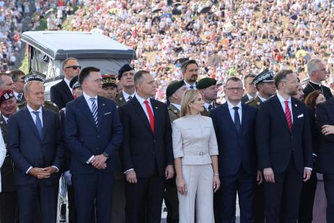 Kilkanaście osób, niektóre w garniturach, a niektóre w mundurach, stoi w trzech rzędach z głowami zwróconymi w stronę defilady. W tle widać tłum ludzi obserwujących defiladę.