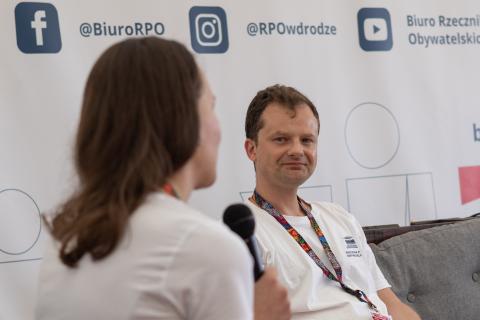 ZRPO Adam Krzywoń siedzi uśmiechnięty i słucha wypowiedzi osoby, która siedzi obok i wypowiada się do mikrofonu