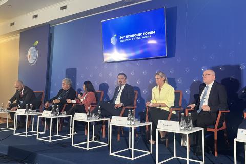 Sześć osób siedzi na krzesłach przy stolikach, na których stoją tabliczki z ich imionami i nazwiskami na tle granatowej ściany z wyświetlaczem, na którym widać napis "34th Economic Forum"