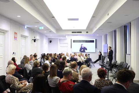 Kilkadziesiąt osób na sali konferencyjnej siedzi i słucha wystąpienia osoby mówiącej za mównicą