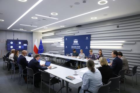 Kilkanaście osób siedzi przy stołach ustawionych w podkowę w sali konferencyjnej, na ścianie duży ekran z widocznymi uczestnikami spotkania łączącymi się przy pomocy oprogramowania do komunikowania się na odległość