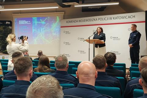 osoba przemawia na stojąco, której słuchają osoby siedzące przed nią