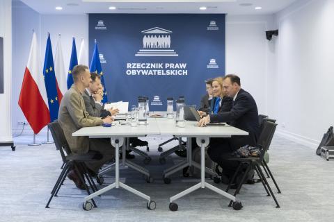 Kilka osób siedzi po dwóch stronach dużego prostokątnego stołu w sali konferencyjnej i rozmawia, w tle na dużej planszy napis "Rzecznik Praw Obywatelskich", a obok stojące flagi Polski i UE