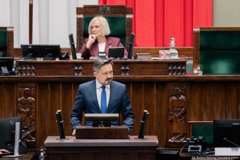 Marcin Wiącek stoi przy mównicy w sali plenarnej Sejmu i wypowiada się. Za nim w fotelu marszałkowskim siedzi wicemarszałkini Dorota Niedziela.
