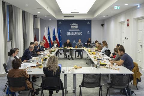 Kilkanaście osób siedzi przy stołach ustawionych w prostokąt na sali konferencyjnej