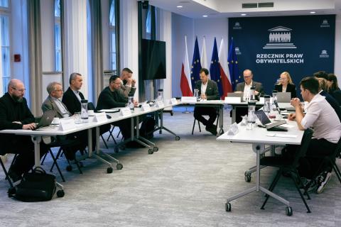 Kilkanaście osób siedzi przy stołach ustawionych w podkowę, za stołami przy ścianie rząd flag Polski i UE