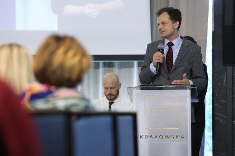 Osoba przemawia na mównicy, za nią widać fragment ekranu, przed nią publiczność siedzi na krzesłach